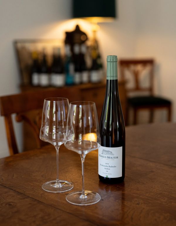 Weinflasche und zwei leere Weinglaeser auf einem Holztisch vor Stuehlen und Flaschen im Hintergrund