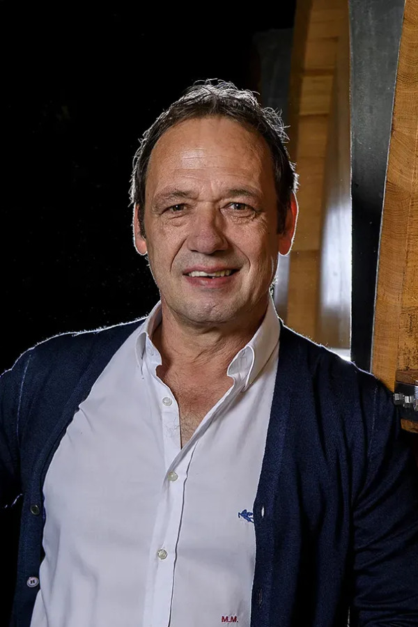 Mann mit weissem Hemd dunkelblauer Strickjacke und Uhr lehnt an Holzfass vor schwarzem Hintergrund